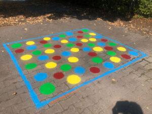 Buntes Twister-Spiel auf einem gepflasterten Platz, umrahmt von blauer Farbe, mit roten, grünen, gelben und blauen Punkten für Freizeitaktivitäten im Freien.