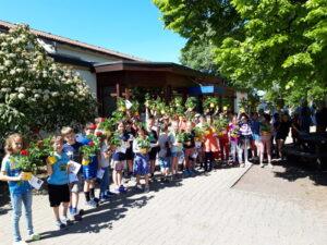 Gruppe von Kindern steht vor einer Schule und hält bunte Blumen in Töpfen. Die Blumen sind ein Geschenk, und die Kinder zeigen stolz ihre Pflanzen. Im Hintergrund sind Bäume und das Schulgebäude sichtbar, bei schönem Wetter.