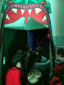 Eine Gruppe von Kindern mit Masken steht vor einem grünen, fantasievollen Eingang mit einem monsterartigen Mund. Sie scheinen auf dem Weg zu einer Veranstaltung oder Attraktion zu sein, während bunte Lichter den Raum erhellen.