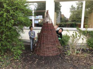 Kinder spielen hinter einem selbstgebauten Weidenhaus im Freien, umgeben von Büschen und einem Fenster im Hintergrund.