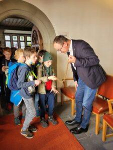 Eine Gruppe von Kindern beobachtet fasziniert einen Mann in Anzug, der eine Kette hält. Die Szene spielt sich in einem historischen Gebäude ab, mit Holztüren und einem einladenden Eingangsbereich.