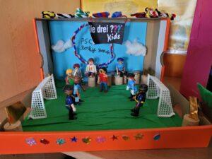 Spielszene mit Playmobil-Figuren auf einem selbstgebastelten Fußballfeld in einer dekorierten Box. Mehrere Figuren stehen im Spiel, zwei Tore sind sichtbar, die Umgebung ist farbenfrohend gestaltet.