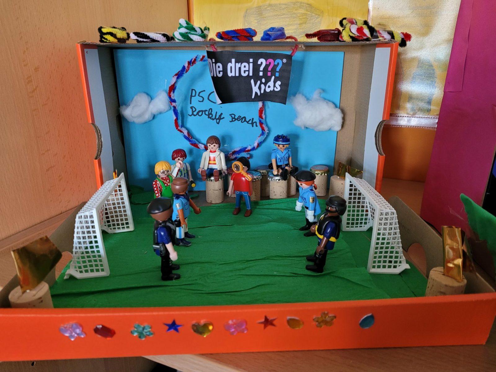 Spielszene mit Playmobil-Figuren auf einem selbstgebastelten Fußballfeld in einer dekorierten Box. Mehrere Figuren stehen im Spiel, zwei Tore sind sichtbar, die Umgebung ist farbenfrohend gestaltet.