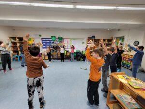 Eine Gruppe von Kindern in einem Klassenzimmer führt eine Bewegungsübung durch, während sie ihre Hände in die Luft heben. Die Umgebung ist freundlich und einladend, mit Regalen voller Materialien und bunten Stühlen im Hintergrund.