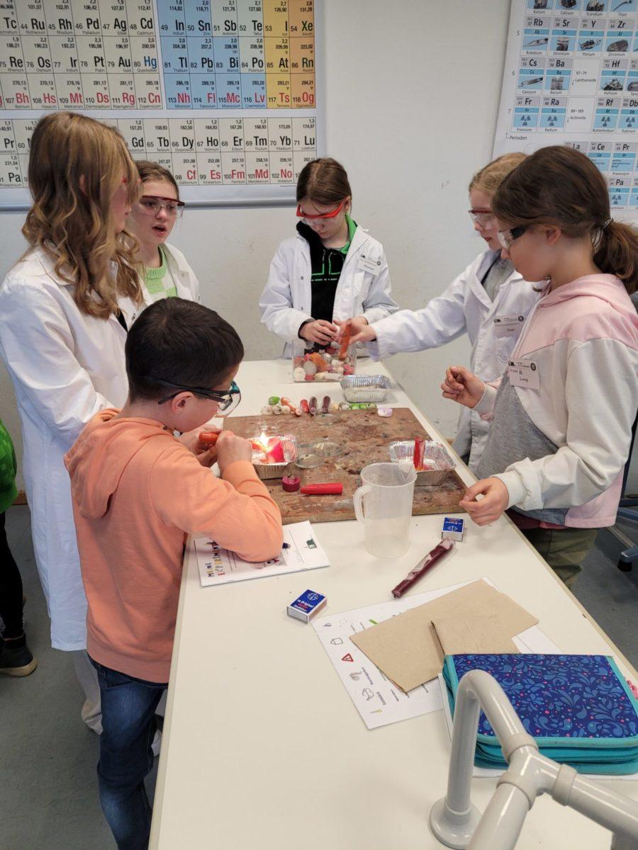 Schüler führen experimentelle Aktivitäten im Chemielabor durch, während sie Schutzbrillen und Laborkittel tragen. Auf dem Tisch sind verschiedene Materialien und Chemikalien sichtbar. Im Hintergrund hängt ein Periodensystem der Elemente.