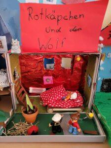 Kreatives Diorama von "Rotkäppchen und der Wolf" mit Figuren, einem Bett, Pflanzen und einer roten Wand. Das Bild zeigt eine fantasievolle Darstellung der Märchenwelt.