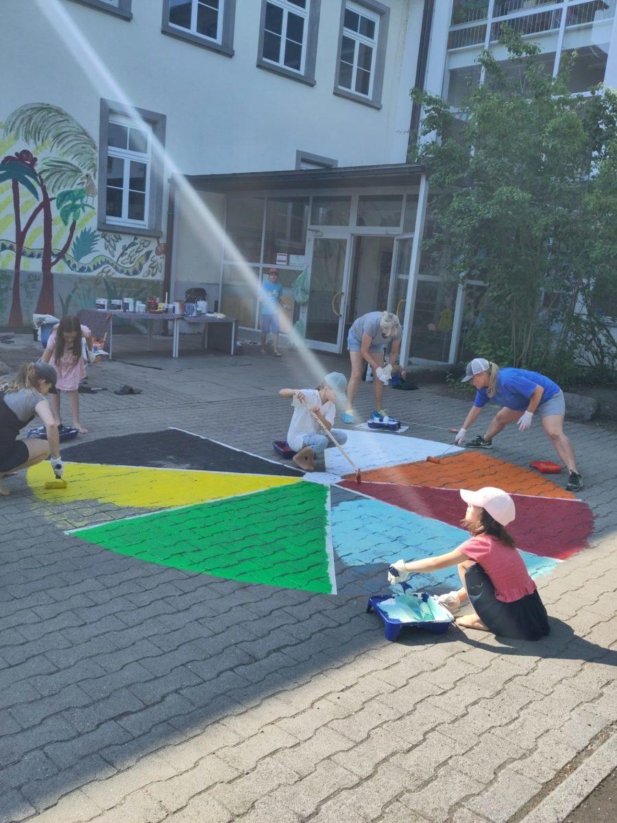 Kinder und Erwachsene malen gemeinsam ein buntes Muster auf dem Boden eines Schulhofs. Im Hintergrund sind Pflanzen und ein Gebäude sichtbar.