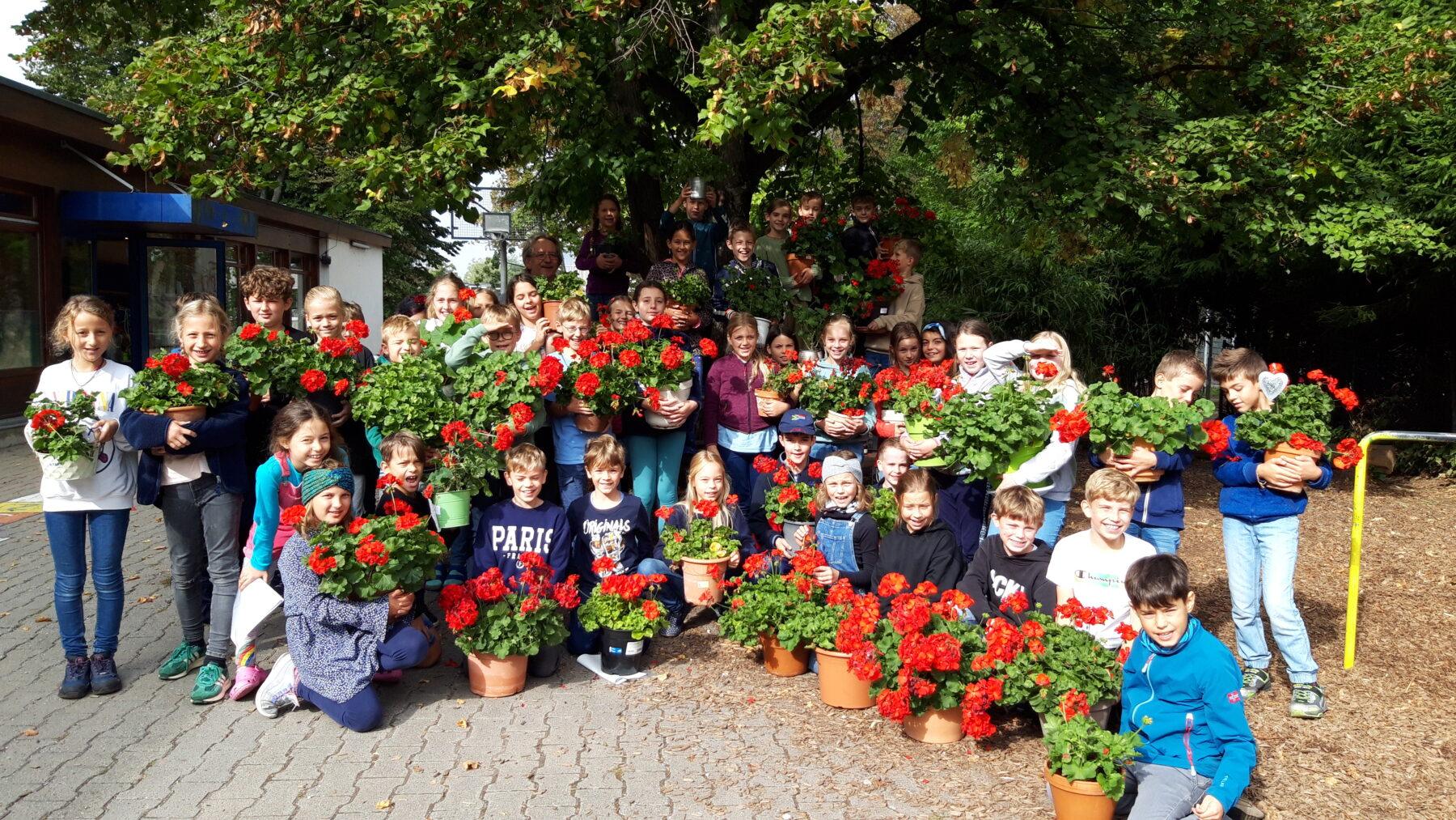Eine Gruppe von Kindern und Erwachsenen steht lächelnd im Freien und hält Töpfe mit roten Geranien. Im Hintergrund ist ein Baum zu sehen. Die Veranstaltung scheint im Rahmen eines Projekts zur Pflanzenpflege oder Blumenschmuckgestaltung stattzufinden.