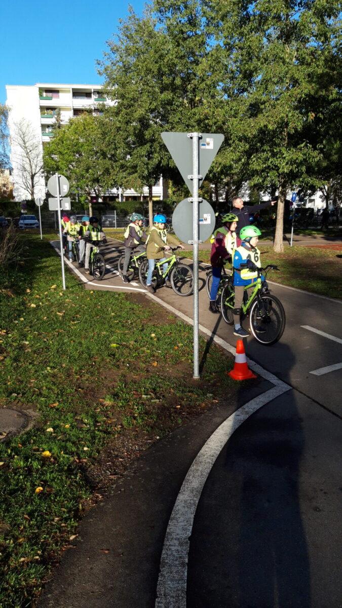 Gruppe von Kindern in Fahrradhelmen, die auf einem Radweg an einer Verkehrszeichen-Kreuzung anhalten. Im Hintergrund sind Bäume und Wohngebäude sichtbar.