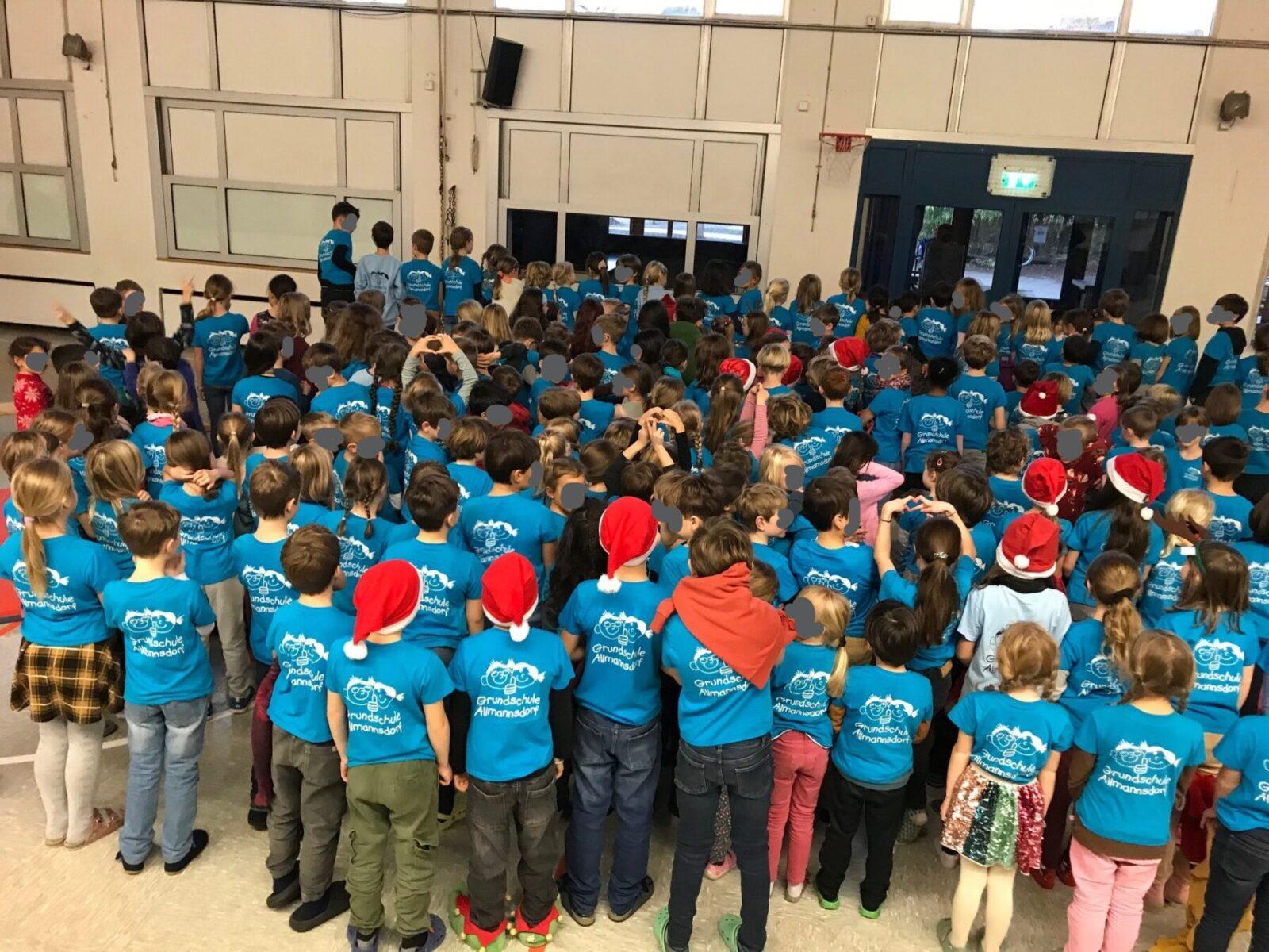 Gruppenschüler der Grundschule Almansdorf in blauen T-Shirts versammeln sich in einer Turnhalle, einige tragen Weihnachtsmützen.