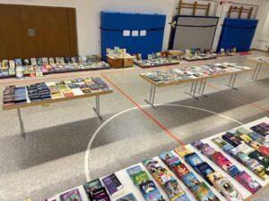 Übersicht über eine Buchausstellung in einer Turnhalle, mit mehreren Tischen, die mit einer Vielzahl von Büchern und Medien zum Thema Kinder- und Jugendliteratur gedeckt sind.