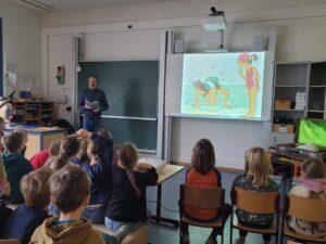 Ein Lehrer liest Kindern in einem Klassenzimmer aus einem Buch vor, während eine farbenfrohe Illustration auf einer Leinwand gezeigt wird. Die Schüler sitzen aufmerksam und zeigen Interesse an der Geschichte.