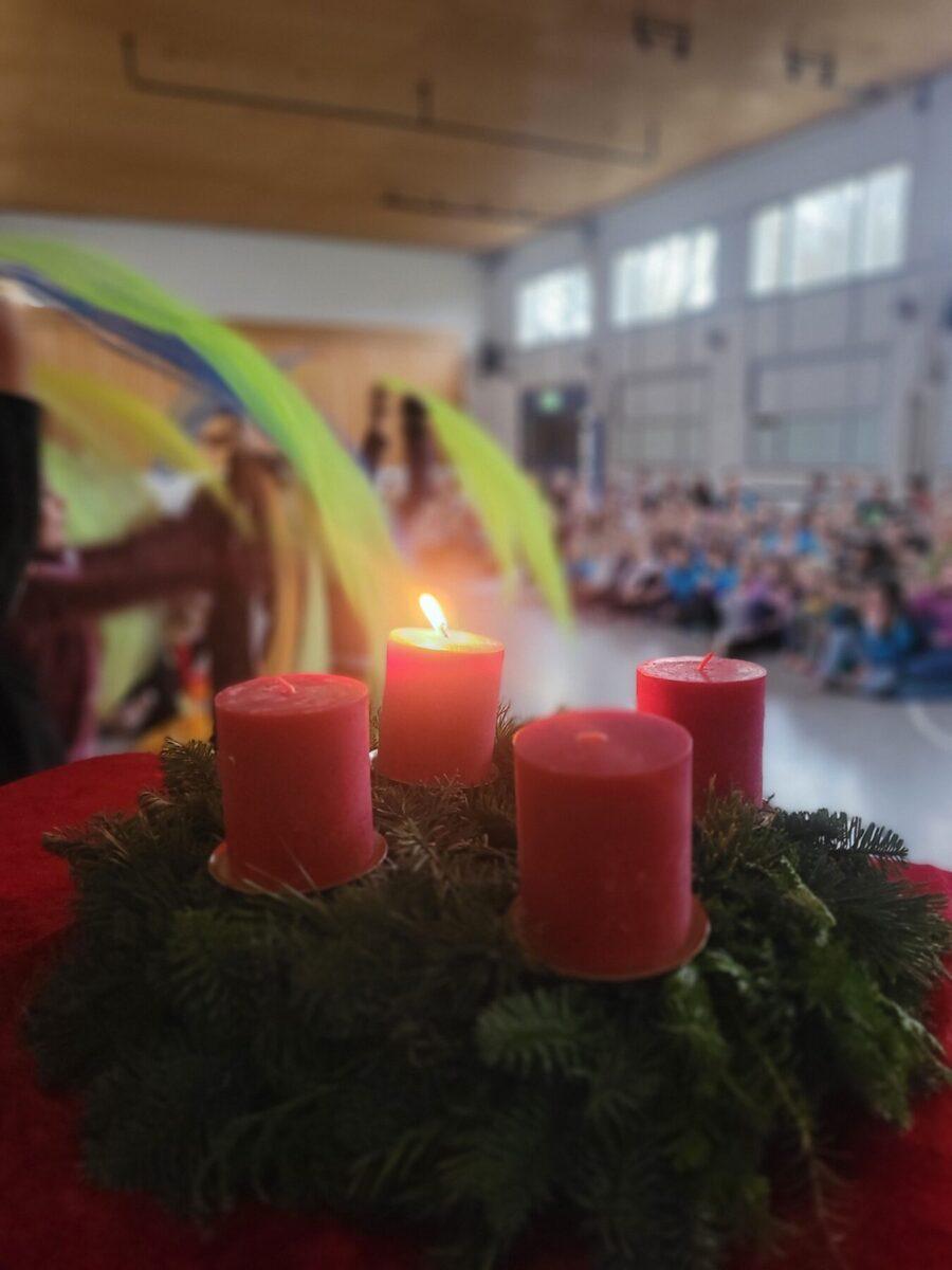 Adventskranz mit roten Kerzen und einer brennenden Kerze im Vordergrund, während im Hintergrund eine festliche Veranstaltung mit Kindern stattfindet.
