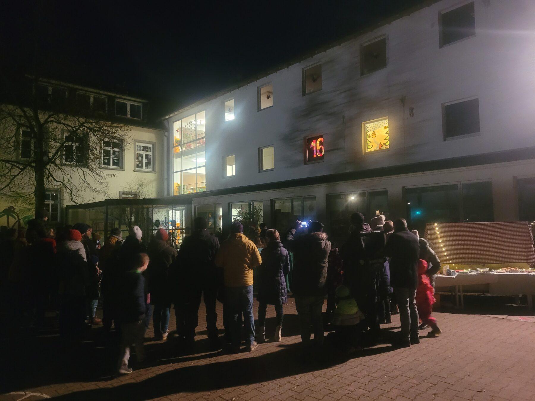 Gruppierung von Menschen vor einem beleuchteten Gebäude bei Nacht, mit einem Adventskalender und festlicher Dekoration. Ein Tisch mit Snacks steht im Vordergrund, während die Teilnehmer auf das Ereignis warten.