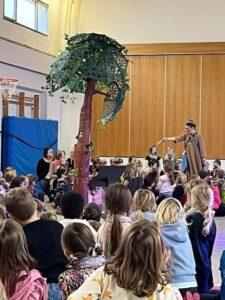 Aufführung für Kinder in einer Turnhalle mit einer Frau, die eine Geschichte erzählt, während die Kinder gebannt zuhören. Im Hintergrund steht ein dekorierter Baum aus Pappe.