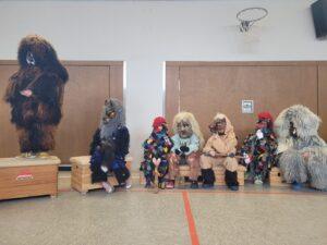 Gruppendarstellung von Personen in fantasievollen Kostümen, die in einer Turnhalle sitzen. Die Kostüme sind tierisch und farbenfroh, inspiriert von traditionellen Masken. Im Hintergrund sind Basketballkörbe und Holzbänke zu sehen.