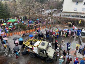 Festliche Versammlung mit vielen bunten Regenschirmen, Menschen in Kostümen und einem historischen Fahrzeug auf einer nassen Straße. Bunte Fahnen und Dekorationen schmücken die Umgebung während einer Feier, möglicherweise ein Karneval oder ein ähnliches Event.