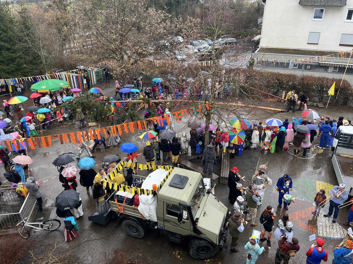 Festliche Versammlung mit vielen bunten Regenschirmen, Menschen in Kostümen und einem historischen Fahrzeug auf einer nassen Straße. Bunte Fahnen und Dekorationen schmücken die Umgebung während einer Feier, möglicherweise ein Karneval oder ein ähnliches Event.