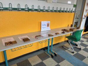 Tisch mit verschiedenen kniffligen Figuren und Informationsblättern in einem Klassenraum. Im Hintergrund eine orangefarbene Wand und eine grüne Stuhl.